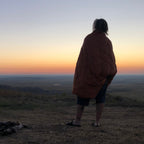 Camper wrapped in Jacks R Better Shenandoah camping quilt watching sunrise over the Badlands from hilltop campsite with stone fire ring