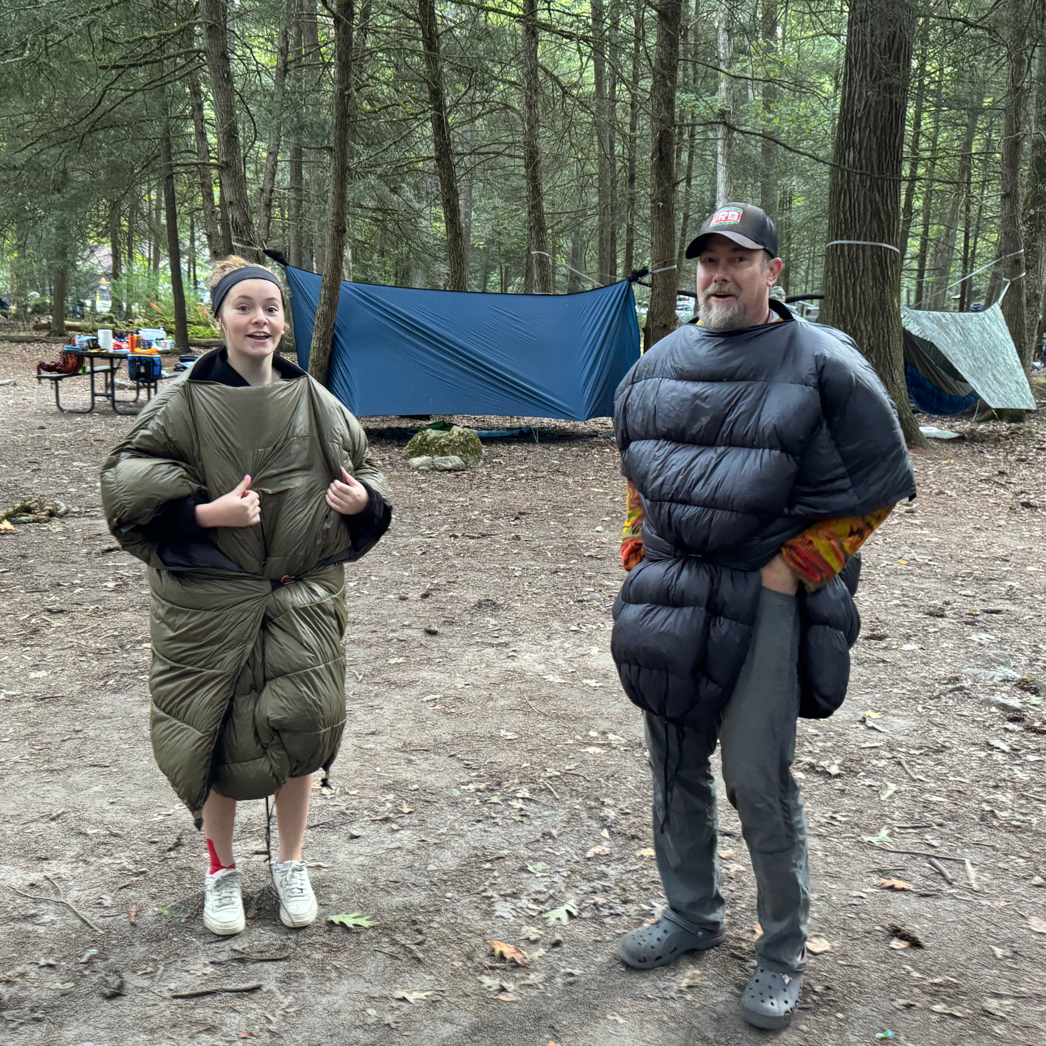 Two campers wearing Jacks R Better Sierra Sniveller 20° wearable down quilts at a wooded campsite